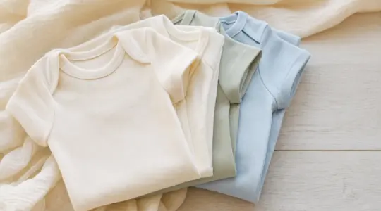 Mehrere gefaltete Baby-Bodys aus Baumwolle in zarten Farben wie Creme, Salbeigrün und Hellblau auf einem hellen Holzuntergrund