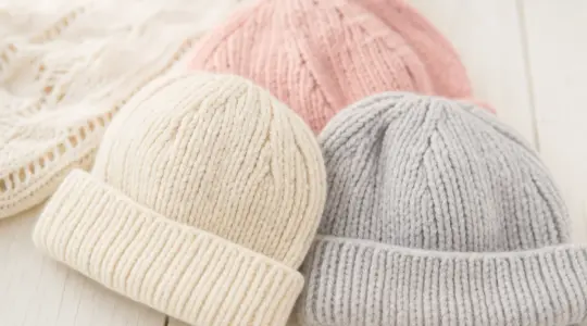 Mehrere weiche gestrickte Baby-Mützen in zarten Farben wie Creme, Hellgrau und Altrosa auf einem hellen Holzuntergrund