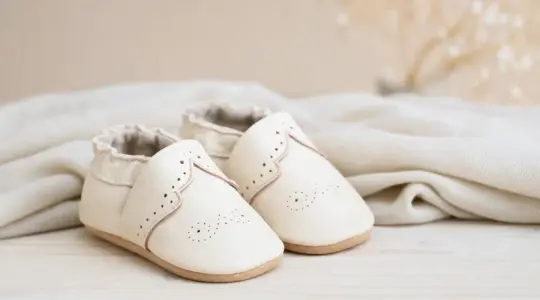 Ein Paar weiche Baby-Krabbelschuhe aus hellem Leder mit zarten Details auf einem hellen Holztisch