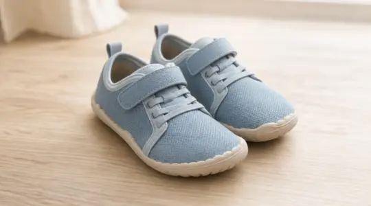 Ein Paar leichte Barfußschuhe für Kinder in zarten Farben mit dünner, flexibler Sohle steht ordentlich auf einem hellen Holzboden