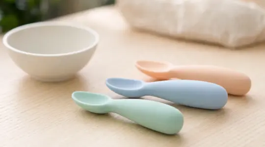 Mehrere weiche Baby-Beikostlöffel aus Silikon mit kurzen Griffen in sanften Farben nebeneinander auf einem hellen Holztisch