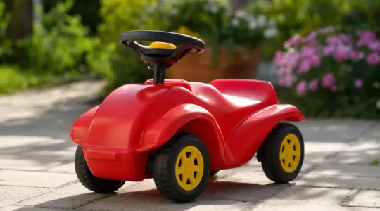 Ein klassisches rotes Kinder-Rutscherauto aus Kunststoff mit gelben Rädern auf einer sonnigen Terrasse im Garten