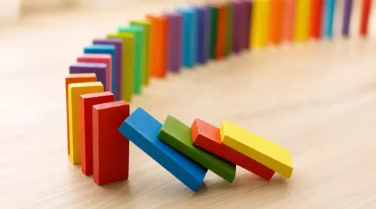 Viele bunte Holz-Dominosteine in einer geschwungenen Kettenreihe aufgestellt, einige fallen um, auf einem hellen Holztisch