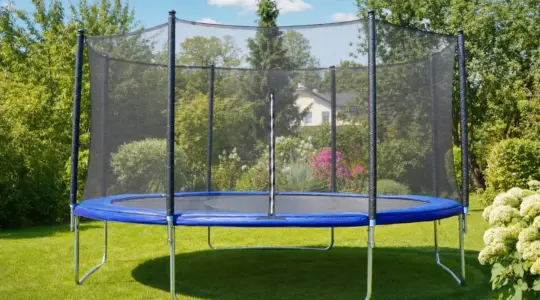 Ein großes Gartentrampolin mit Sicherheitsnetz steht auf einem grünen Rasen bei Sonnenschein bereit