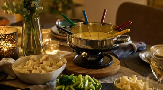 Ein Fondue-Set mit Topf und mehreren bunten Gabeln steht auf einem gedeckten Tisch, daneben Schälchen mit Zutaten