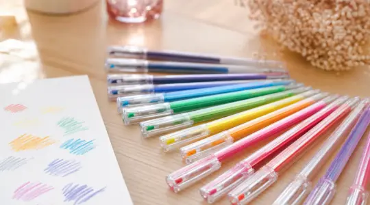 Ein Set bunter Gelstifte in vielen leuchtenden Farben liegt aufgefächert auf einem hellen Tisch neben einem Blatt Papier