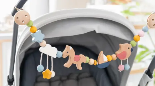Eine bunte Kinderwagenkette aus Holzperlen und kleinen Holzfiguren in fröhlichen Farben dekorativ aufgehängt vor einem hellen Hintergrund