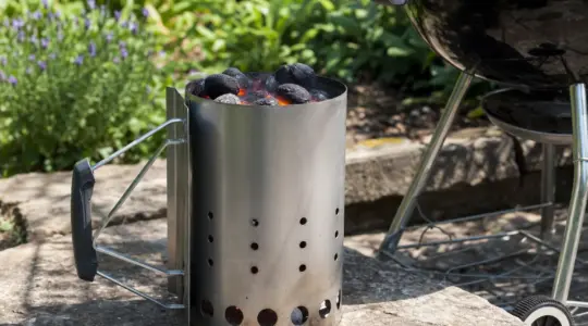 Ein silberner Anzündkamin mit glühender Grillkohle steht neben einem Grill