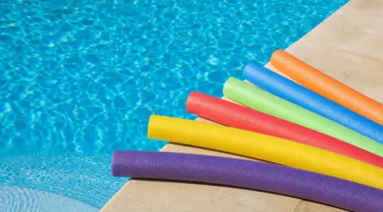 Mehrere bunte Poolnudeln aus Schaumstoff am Rand eines Swimmingpools