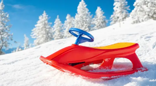 Ein bunter Schneerutscher aus Kunststoff mit Lenker steht im frischen Schnee an einem sonnigen Winterhang bereit