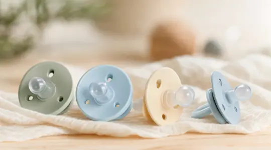 Mehrere moderne Baby-Schnuller mit weichem Silikon-Sauger in zarten Farben auf einem hellen Holztisch mit Mulltuch