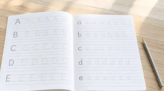 Ein aufgeschlagenes Schreiblernheft mit Lineatur und vorgedruckten Übungsbuchstaben liegt mit einem Bleistift auf einem Tisch